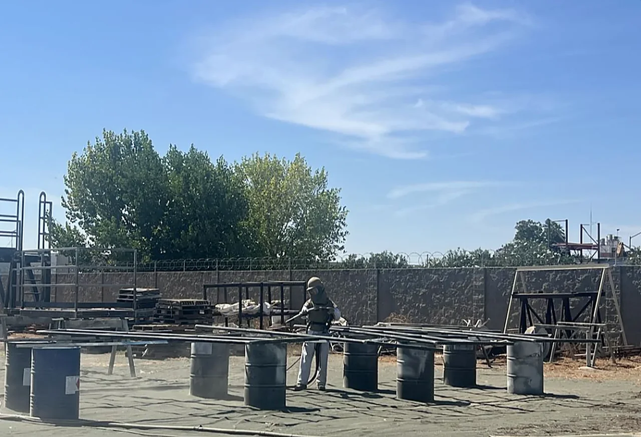 Gold Star Painting technician in a blast hood sandblasting steel railing sections at the Modesto yard